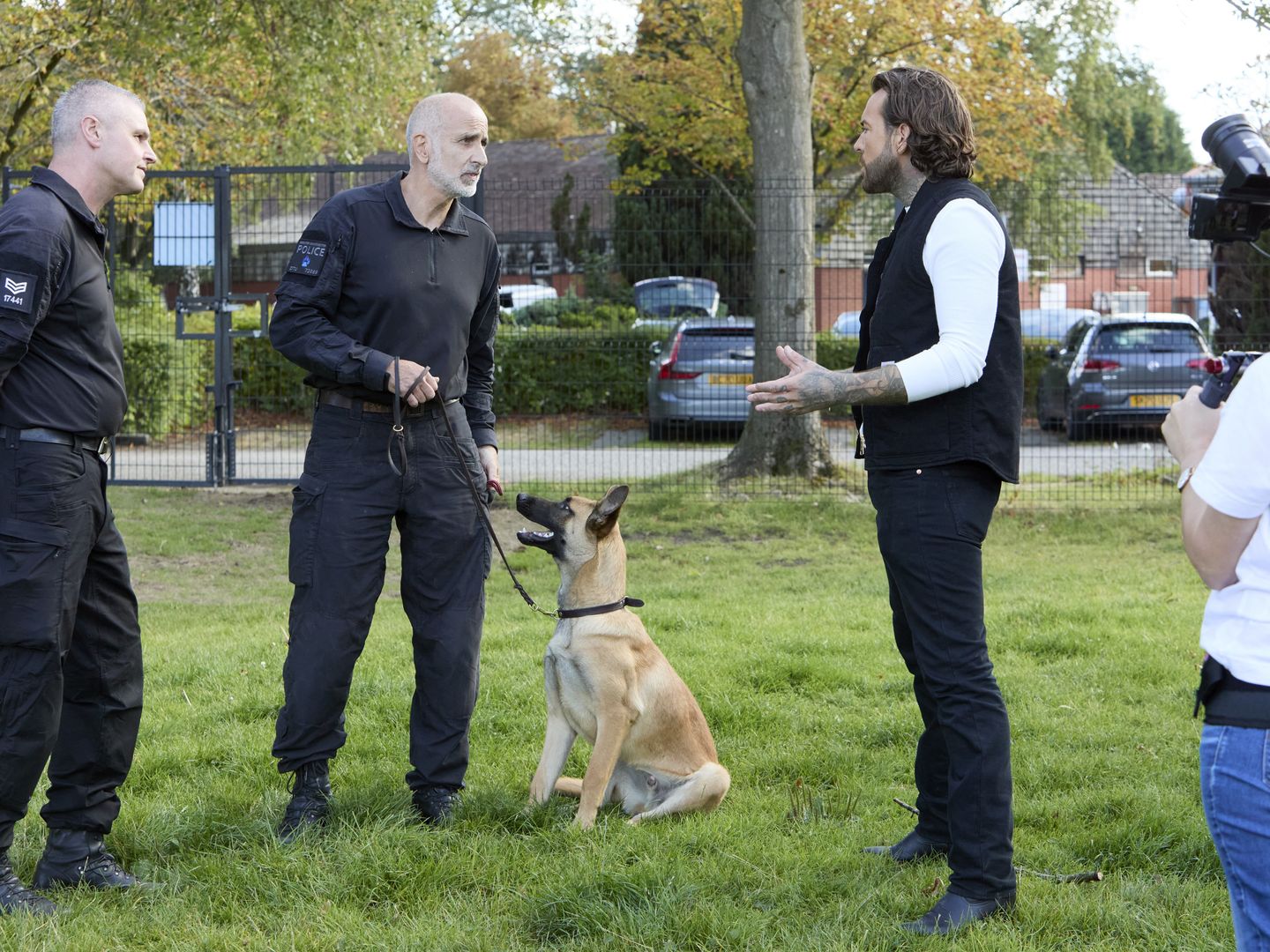 Pete Wicks standing outside talking to two male police officers with one holding a lead with a German Shepherd sitting and looking up next to him