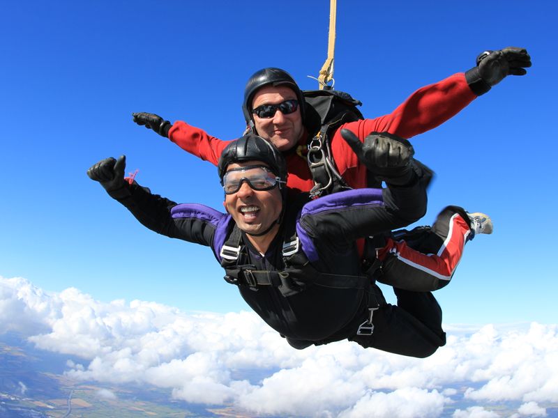 Two men tandem skydiving together free falling in the sky, looking at smiling at the camera